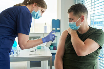 Female doctor spraying disinfectant on arm of male patient before giving COVID-19 vaccine at clinic