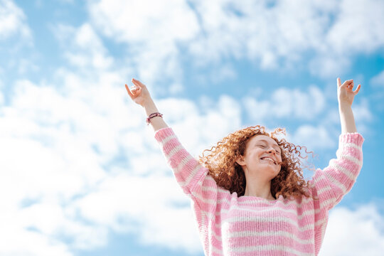 Happy redhead woman with arms raised dancing under sky