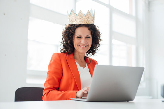 Smiling female professional wearing crown while using laptop at office