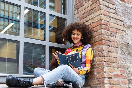 Smiling young woman reading book while sitting by window at university