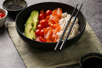 Poke bowl - black bowl with white rice, smoked salmon, cherry tomatoes and avocado, sesame seeds, soy sauce and chopsticks on a grey concrete surface, close-up