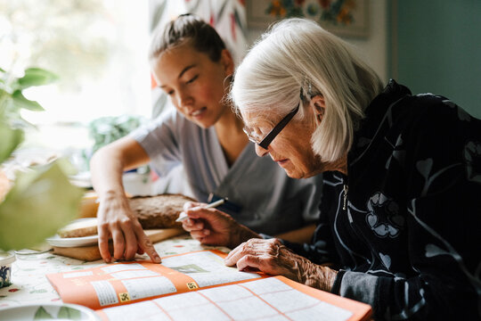 Female nurse solving crossword puzzle with senior woman while sitting in kitchen at home