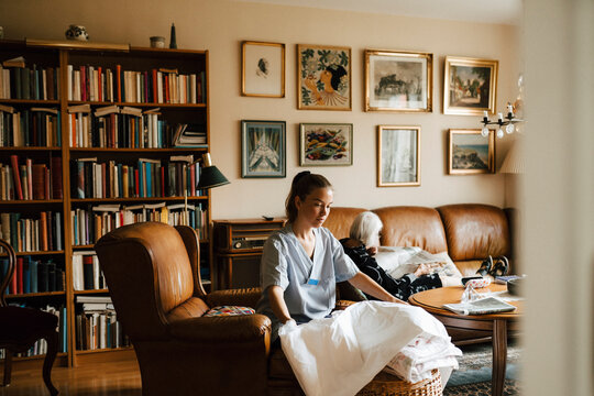 Female nurse folding blanket while senior woman reading newspaper in living room