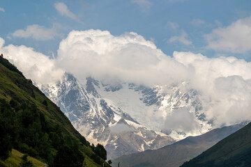 A panoramic view on the snow-capped peak of of Jangi-Tau(Dzhangi-Tau) in the Greater Caucasus Mountain Range in Georgia, Svaneti Region. Hills with lush pastures, peaks covered in clouds.