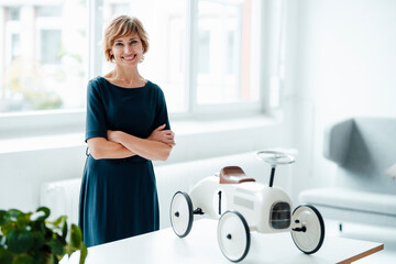 Female business professional with arms crossed standing by toy car in office