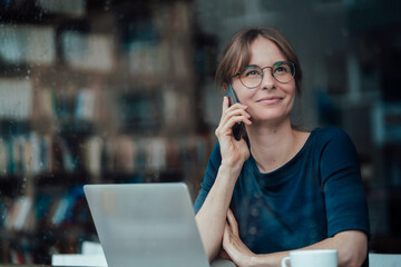 Smiling female professional looking away talking on smart phone in cafe seen through glass window