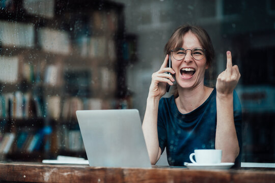 Cheerful businesswoman talking on smart phone while showing middle finger through cafe window