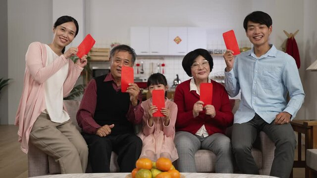 happy family of mixed generation smiling at camera with thumb and victory hand gestures while taking burst photos with red envelopes lucky money at home on Chinese new year.  translation: fortune