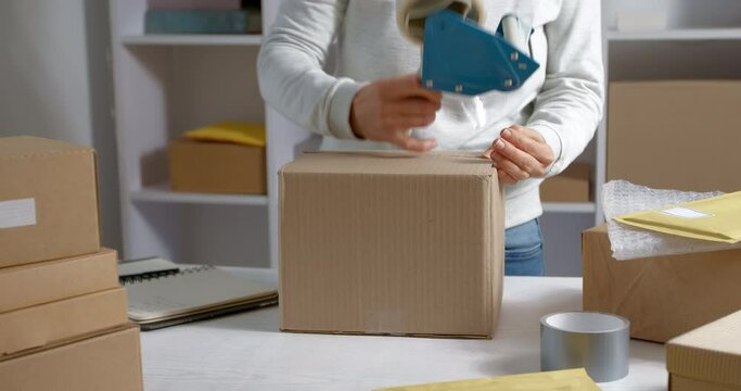 Warehouse worker packs a box of goods for courier delivery. A woman is packing a cardboard parcel with a dispenser with scotch tape.