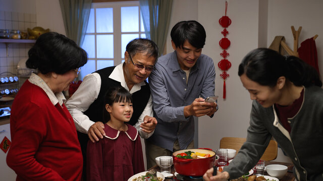 excited asian male father taking photos of family getting together for chinese new year's dinner by dining table with mobile phone while young mother is cooking hot pot