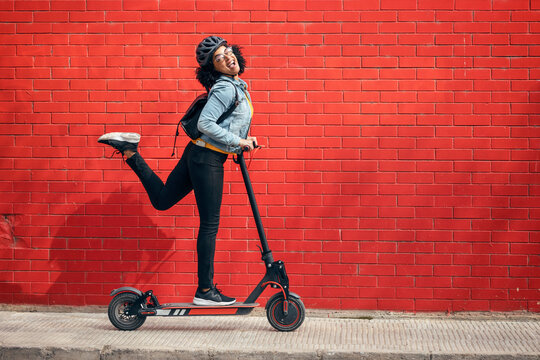 Carefree young woman riding electric push scooter by red brick wall