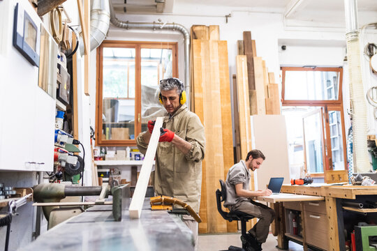 Mature male carpenter working on wood while coworker writing in workshop