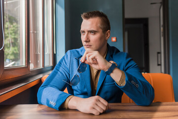 A brooding, stylish guy businessman of Caucasian attractive appearance holds glasses in his hands and looks out the window sitting at a table in a cafe