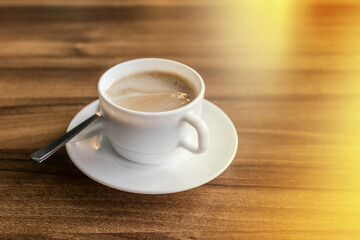 A cup of hot coffee on a saucer with a spoon stands on a wooden background of a table in a cafe