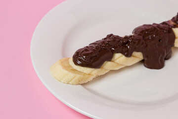 Banana in chocolate on a pink background. Banana slices are drizzled with melted chocolate on a white plate. Fruit dessert