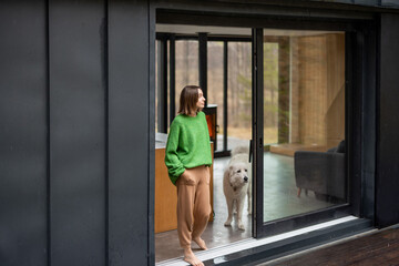 Young woman with her dog at modern house on nature. Girl in green sweater standing near the window...