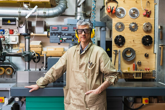 Mature male craftsperson with hand in pocket standing by machinery at workshop