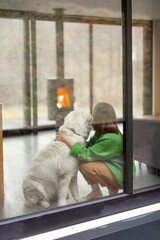 Young woman with her dog at modern house on nature. Girl in green sweater sitting near the window and look on fireplace. Rear view through the window. Concept of comfort and happy modern life