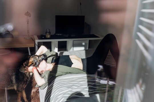 Woman kissing German shepherd dog while lying on bed seen from window