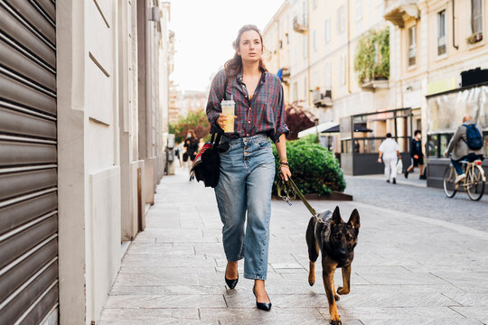 Woman holding drink while walking with German shepherd dog on footpath in city