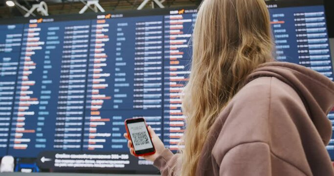 Passenger in the airport looking at QR code on the screen of smartphone mobile phone. Woman travelling with certificate of vaccination from COVID-19. Flights schedule behind