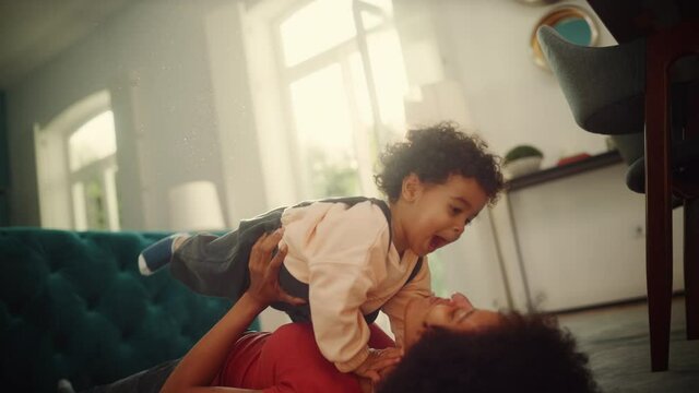 Beautiful African American Mother Playing with Adorable Baby Boy at Home on Living Room Floor. Cheerful Mom Nurturing a Child. Happy Son Raised in the Air. Concept of Childhood, New Life, Parenthood.