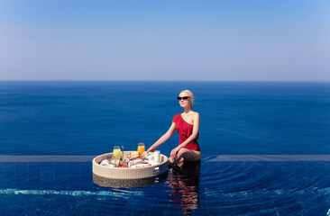 Beautiful blonde caucasian girl in a red swimsuit having breakfast in the swimming pool overlooking the sea.