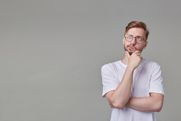 studio portrait of young bearded man wears white t-shirt and spectacles, looks aside with thoughtful facial expression, touching his chin. isolated over gray background.