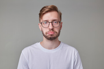 studio portrait of young bearded man wears white t-shirt and glasses looks directly into camera, curving lip with sad facial expression isolated over gray background.