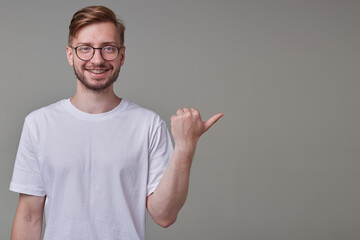 studio portrait of young bearded man wears white t-shirt point with a finger aside at copy space, smiles broadly and feels happy isolated over gray background.