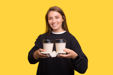 Young kind woman is holding and passing you a pair of to go coffee cups over yellow background.