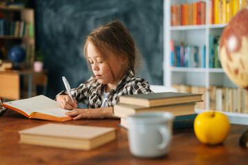 Side view of serious focused pupil kid school girl doing homework writing in notebook sitting at table with paper book. Portrait elementary child schoolgirl studying at home, selective focus.