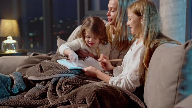 Caucasian mother resting with her two daughters on cozy sofa and drawing in albums with colorful pencils. Leisure time at home of happy family. 