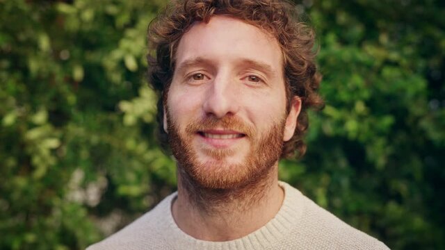Close Up Portrait of a Happy Young Adult Male with, Brown Eyes, Curly Ginger Hair and Beard Posing for Camera. Handsome Diverse Caucasian Male Smiling on Green Nature Background.