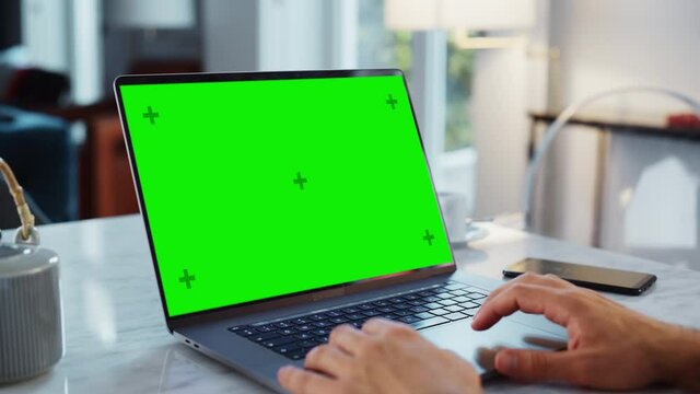 Green Screen Mock Up Display on a Laptop Computer. Close Up on Person's Hands Working from Modern Home, Using Touch Pad, Scrolling Content. Smartphone Lies Next to Computer.