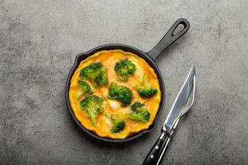 Healthy vegetarian egg omelette with green broccoli in black cast iron frying pan with fork and knife top view on gray stone concrete background table, diet and healthy clean eating concept 