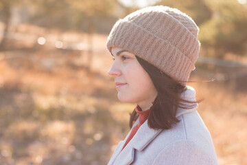 A serious girl in a knitted hat stands against a background and a coat against the backdrop of an autumn pale park landscape.