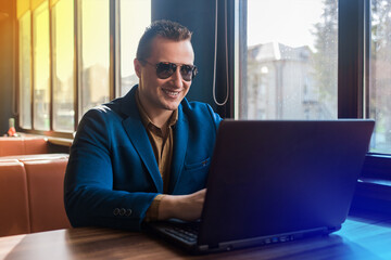 A business man businessman a stylish smiling positive portrait of Caucasian appearance in a blue jacket and sunglasses works in a laptop or computer, sitting at a table by the window in a cafe