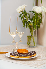 Lighted Hanukkah with one candle and shamash on the first day of Hanukkah on the festive table next to donuts and a vase of flowers.
