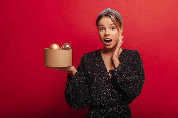 Beautiful young fair-skinned girl looking surprised opening mouth to camera on red background. She holding small box with Christmas tree decorations. Wears shiny black party dress. Celebration concept