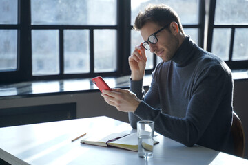 Attractive European male entrepreneur sitting at office table with mobile phone in hand