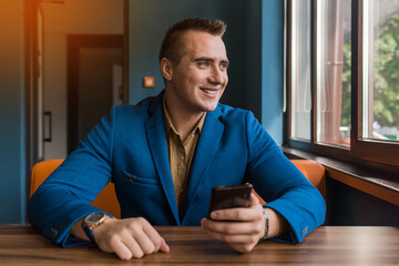 A smiling stylish, young businessman of Caucasian appearance portrait in a jacket and shirt sits at a table in a mobile phone and looks out the window