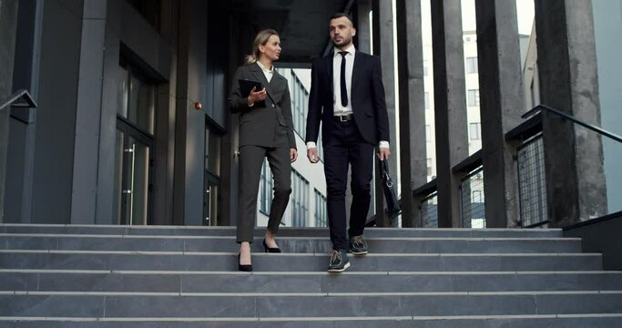 Good looking young male and female business colleagues walking the steps down from the big office building, talking to each other and discussing
