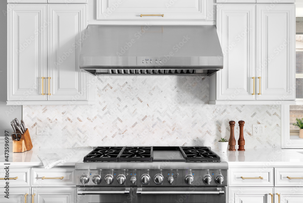 Wall mural A detail shot of a beautiful white kitchen's stainless steel stove, hood, granite counter tops, and a herringbone marble tiled back splash.