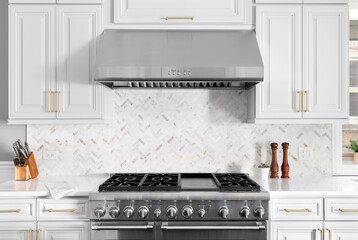 A detail shot of a beautiful white kitchen's stainless steel stove, hood, granite counter tops, and...