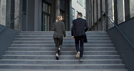 Back view of the successful young businessman and businesswoman stepping up the steps outside the big urban office building