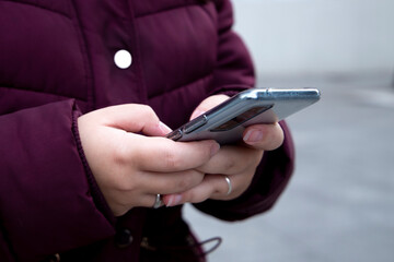 Young girl with a mobile device in her hands typing, wears maroon winter clothes while on the street.