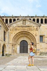 Horizontal view of young woman with backpack sightseeing. Panoramic view of woman looking at the ancient cathedral of Santillana del Mar. Travel and holidays concept in european cities.