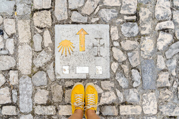 Top view of unrecognizable woman feet hiking the spanish Saint james way. Horizontal view of woman with yellow shoes isolated stepping on Saint James pilgrim sign. Travel and people background.