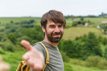 Front view of caucasian man reaching the hand to the camera. Horizontal view of backpacker traveling outdoors with travel destination in the background. People and travel concept.